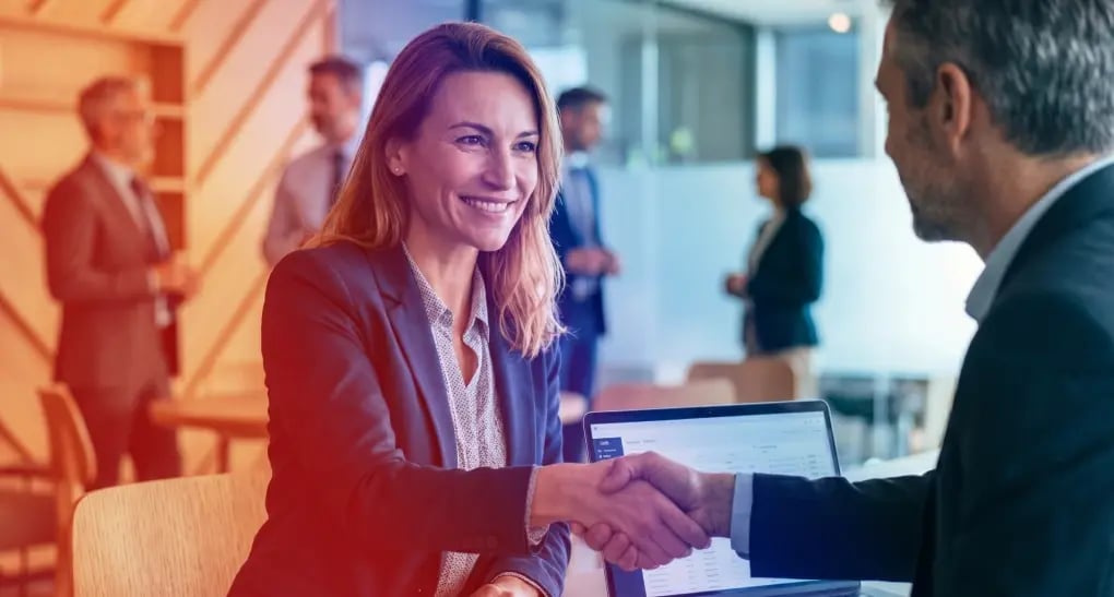 A smiling corporate technology consultant in a blue blazer shakes hands with an enterprise business client next to an open laptop during a strategic consulting engagement.