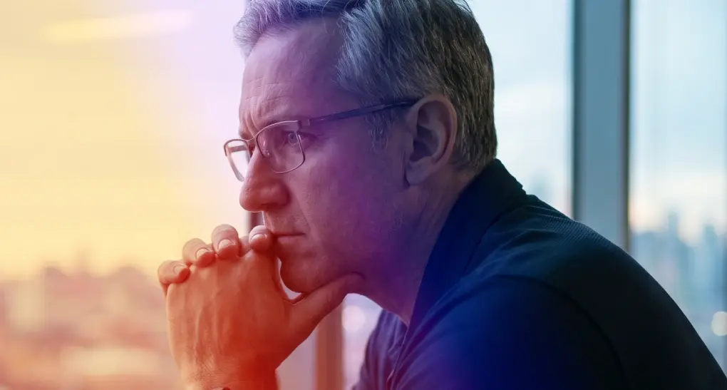 A pensive male technology executive with glasses rests his chin on his hands while staring out an office window, reflecting on engineering delivery bottlenecks.