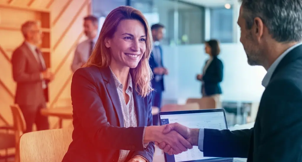 A smiling corporate technology consultant in a blue blazer shakes hands with an enterprise business client next to an open laptop during a strategic consulting engagement.