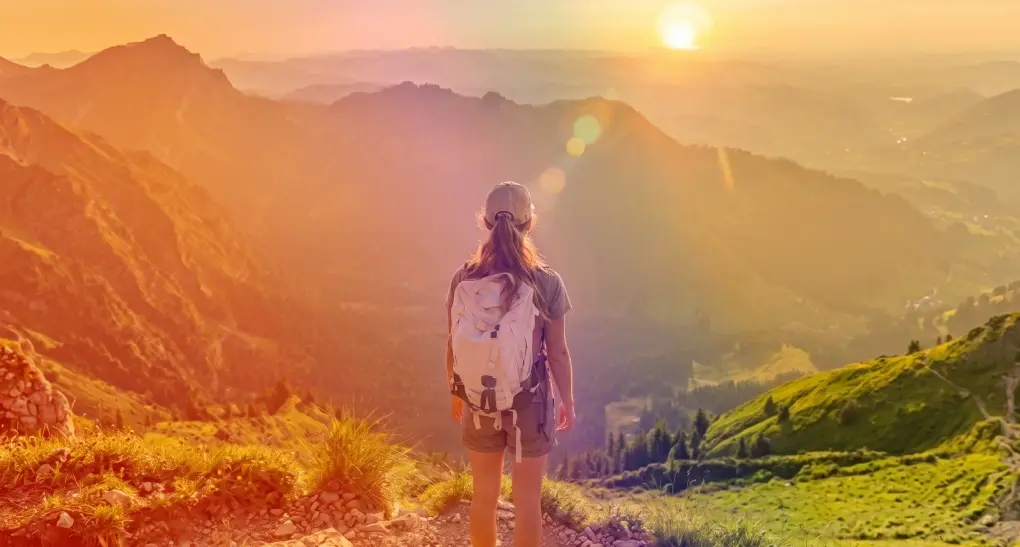 Rear view of a hiker with a white backpack standing on a grassy ridge looking out over a wide mountain valley landscape, symbolizing navigation and strategic guidance.