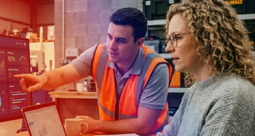 A female UX researcher in a blazer analyzes user research data on a laptop alongside an industrial operations worker in a safety vest during a field discovery session.